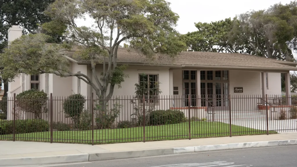 The single-story Minerva Club clubhouse behind a black iron fence and old trees in Santa Maria