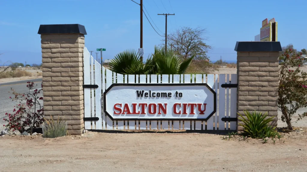 A white Welcome to Salton City sign between two stone pillars, with palms and desert behind